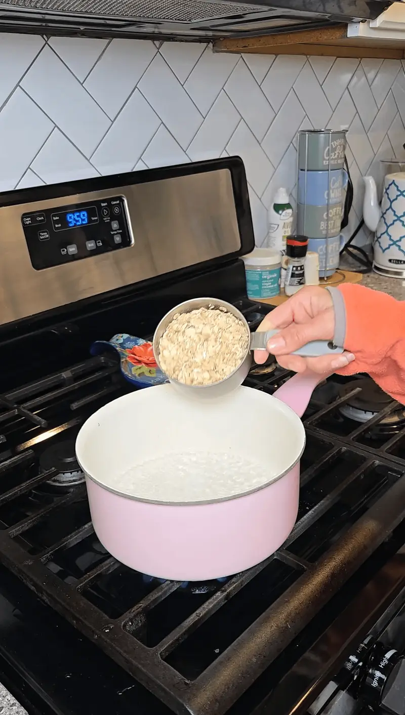 Tipping a measuring cup of rolled oats into a pink saucepan of simmering water
