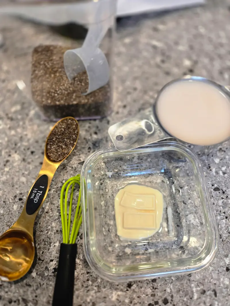 The white chocolate in a glass dish beside measuring spoons of chia and cocoa